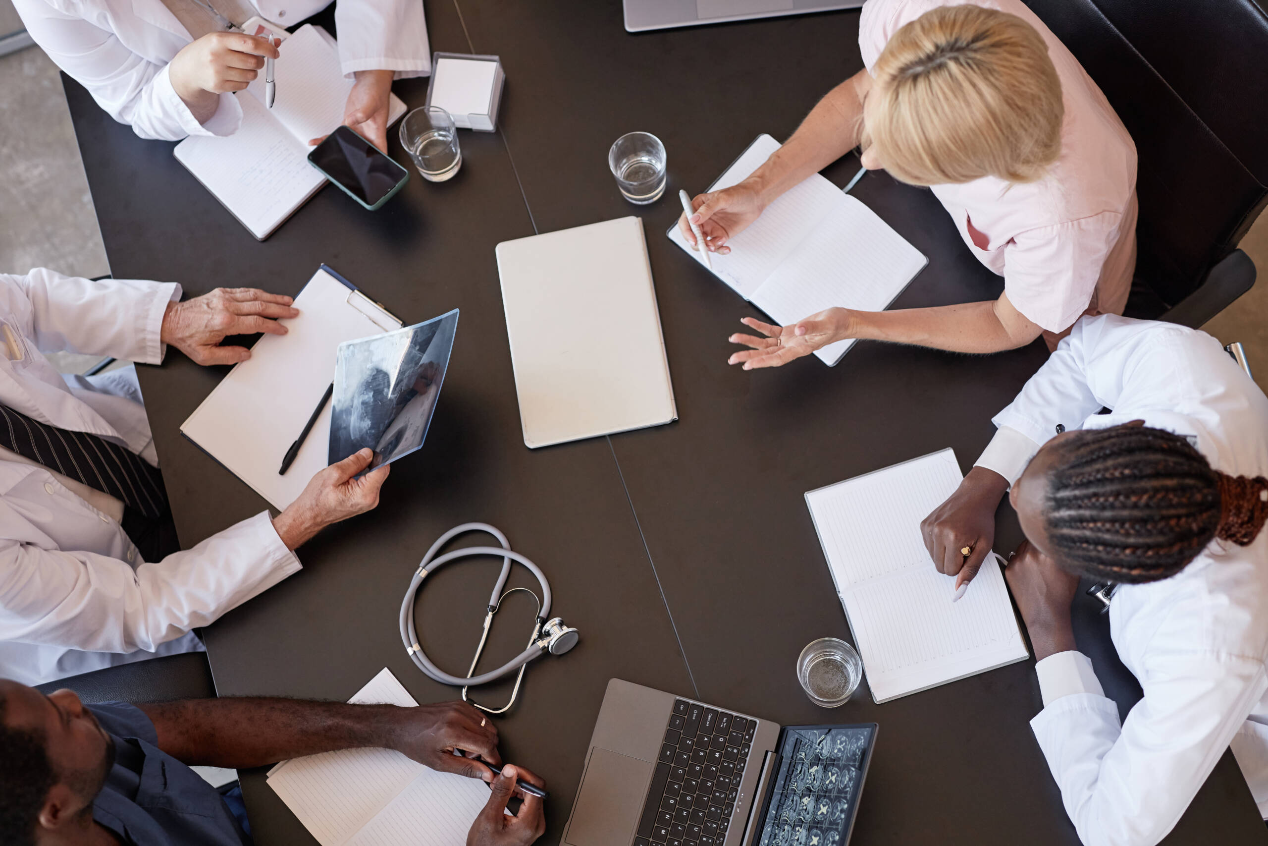 Top view of woman passing CT image of patients skull to chief physician discussing X ray examination results with colleagues, while sitting at table with laptops during meeting at medical office