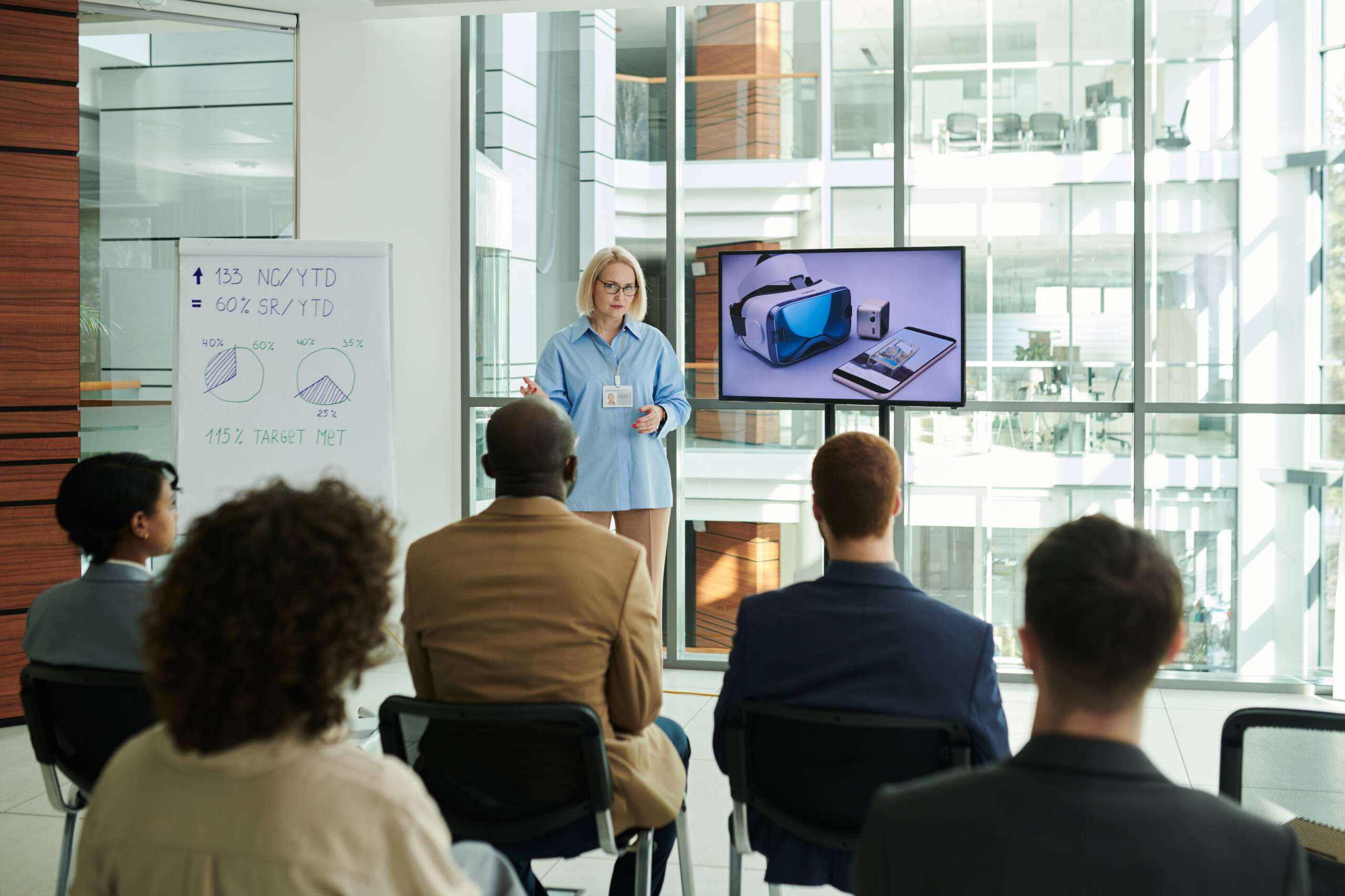 Rear view of young audience listening to mature female coach standing by interactive board with VR headset and smartphone at seminar